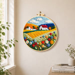 Blooming flower field and red roof cottage needle felt wall art displayed in a bright home interior