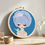 Blue haired girl with bird punch needle artwork displayed on a table