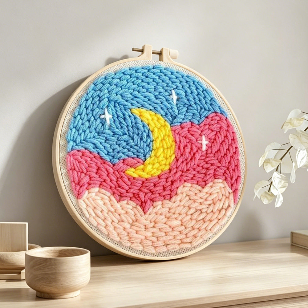 Crescent moon over pink and blue cloud sky punch needle artwork displayed on a table