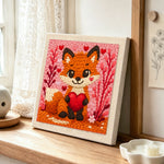 Fox holding a red heart punch needle artwork displayed on a table