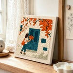 Girl with orange leaves at blue door punch needle artwork displayed on a table