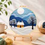 Finished minimalist blue mountains and snowy house needle felting displayed on a wooden easel