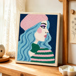 Pastel blue haired woman in pink beret punch needle artwork displayed on a table