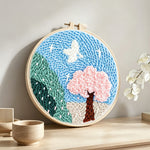 Pink blossom tree and green mountains punch needle artwork displayed on a table