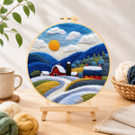 Finished red barn and snowy blue hills needle felting displayed on a wooden easel