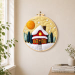 Red cottage under snowy yellow sky needle felt wall art displayed in a bright home interior