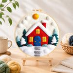 Finished red winter house with blue door needle felting displayed on a wooden easel