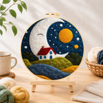 Finished smiling giant moon and little red cottage needle felting displayed on a wooden easel