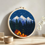 Snow-capped mountains, dark forest and campfire punch needle artwork displayed on a table
