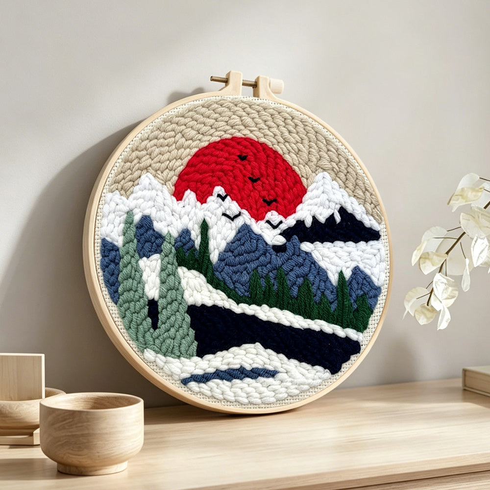 Snowy mountain landscape with pine trees and red sun punch needle artwork displayed on a table