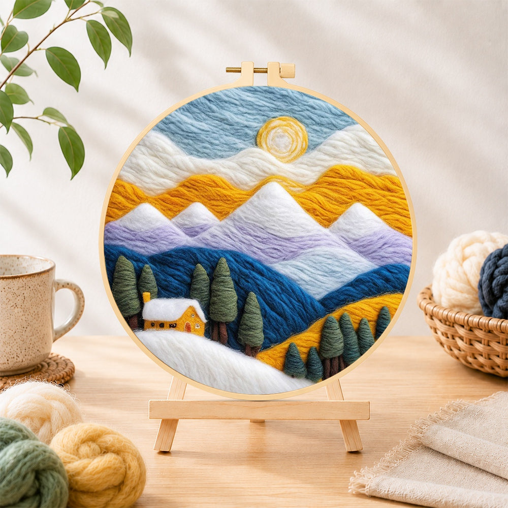 Finished snowy peaks and yellow cottage with trees needle felting displayed on a wooden easel