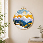 Snowy peaks and yellow cottage with trees needle felt wall art displayed in a bright home interior