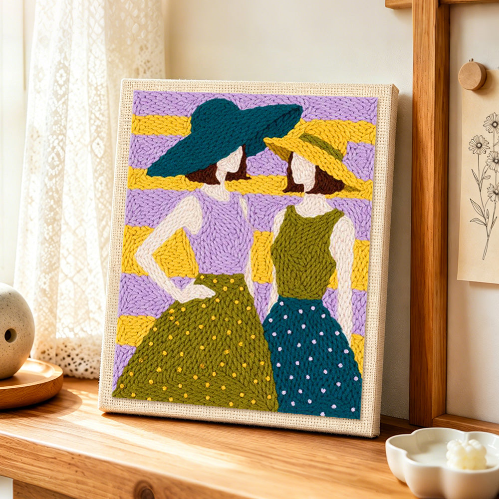 Two women in sun hats punch needle artwork displayed on a table