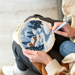 Creating handmade winter cabin scene with snowy pine trees and blue river punch needle artwork
