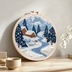 Winter cabin scene with snowy pine trees and blue river punch needle artwork displayed on a table